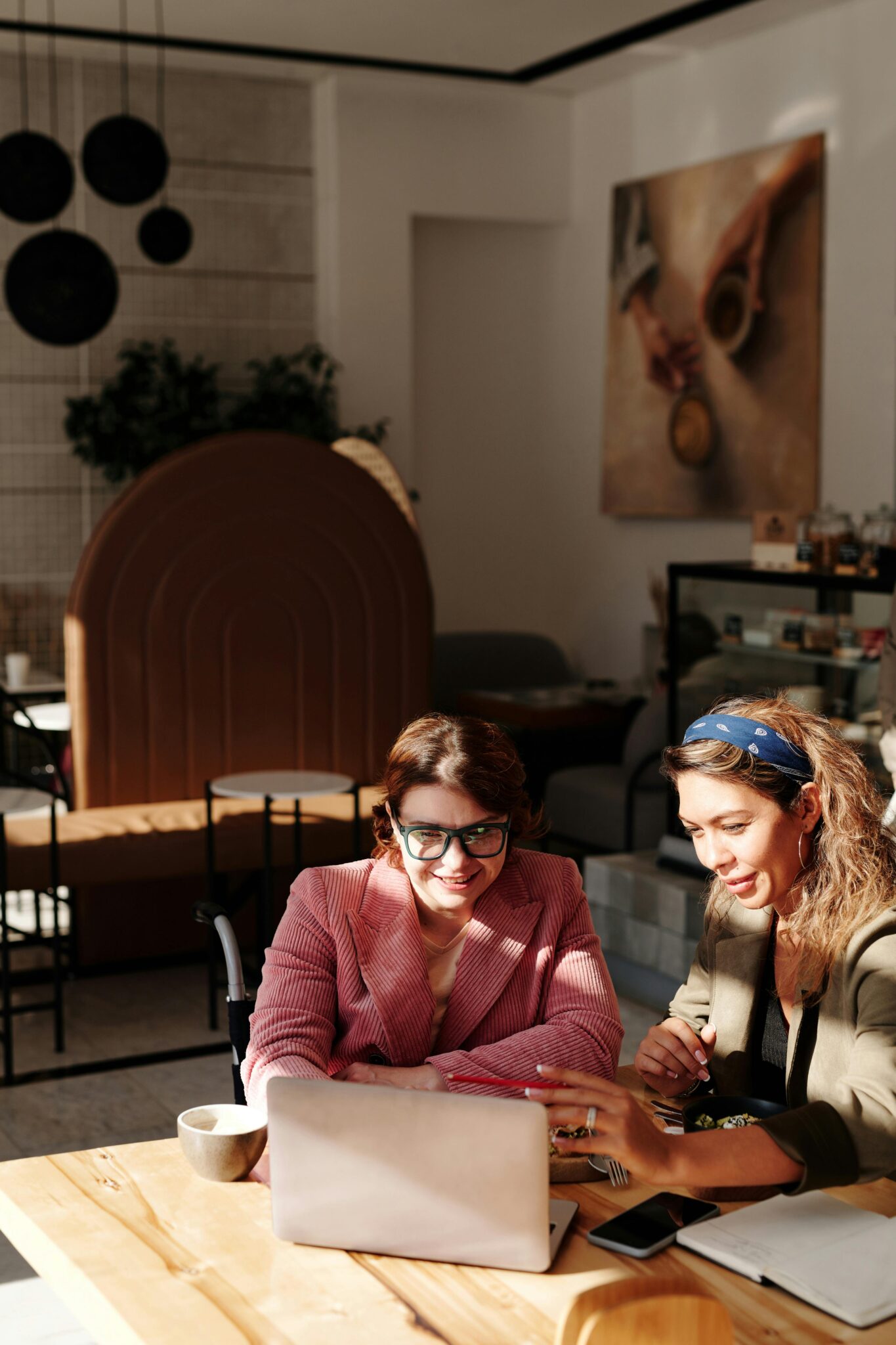 Two women working together in a bright café with laptops and coffee cups.